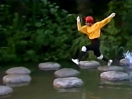 A contestant in a yellow top and black trousers leaps across stepping stones in a pond.