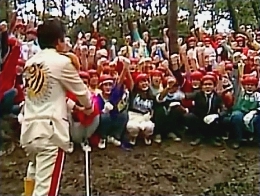 General Tani faces a large group of contestants squatting on the ground in a wooded area