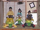 Three pairs of parent and child players in an earthquake simulator decorated like a Japanese house. The parents wear turtle shells and crouch on their hands and knees while their children, wearing wigs and holding wooden chests, balance on their backs.