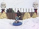 A man sitting on a round blue sledge is pushed down a snowy hill